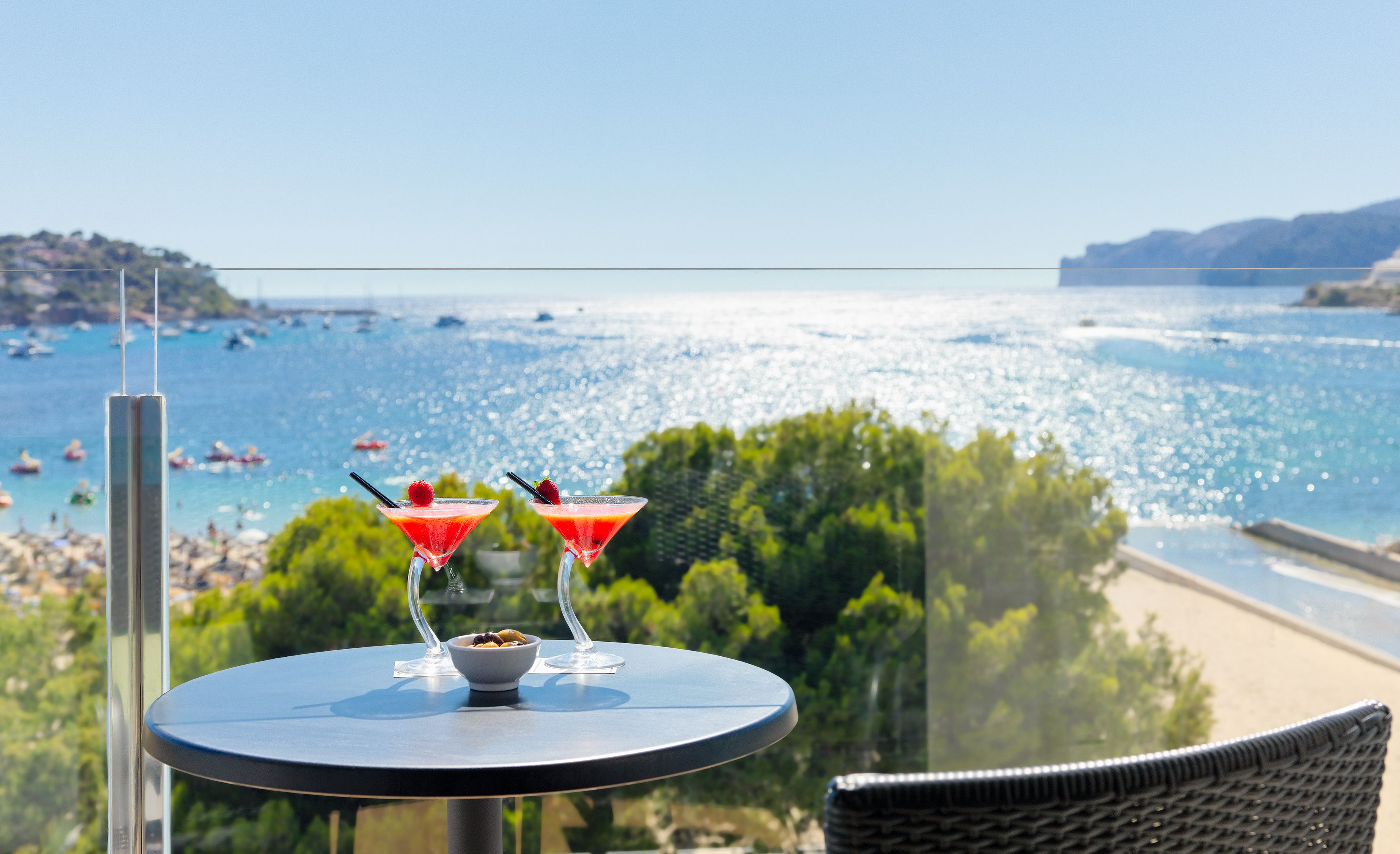 A balcony with two martini glasses and a bowl of fruit, overlooking a beach and the sea.