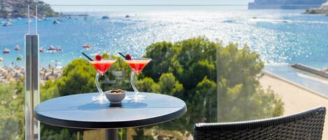 A balcony with two martini glasses and a bowl of fruit, overlooking a beach and the sea.