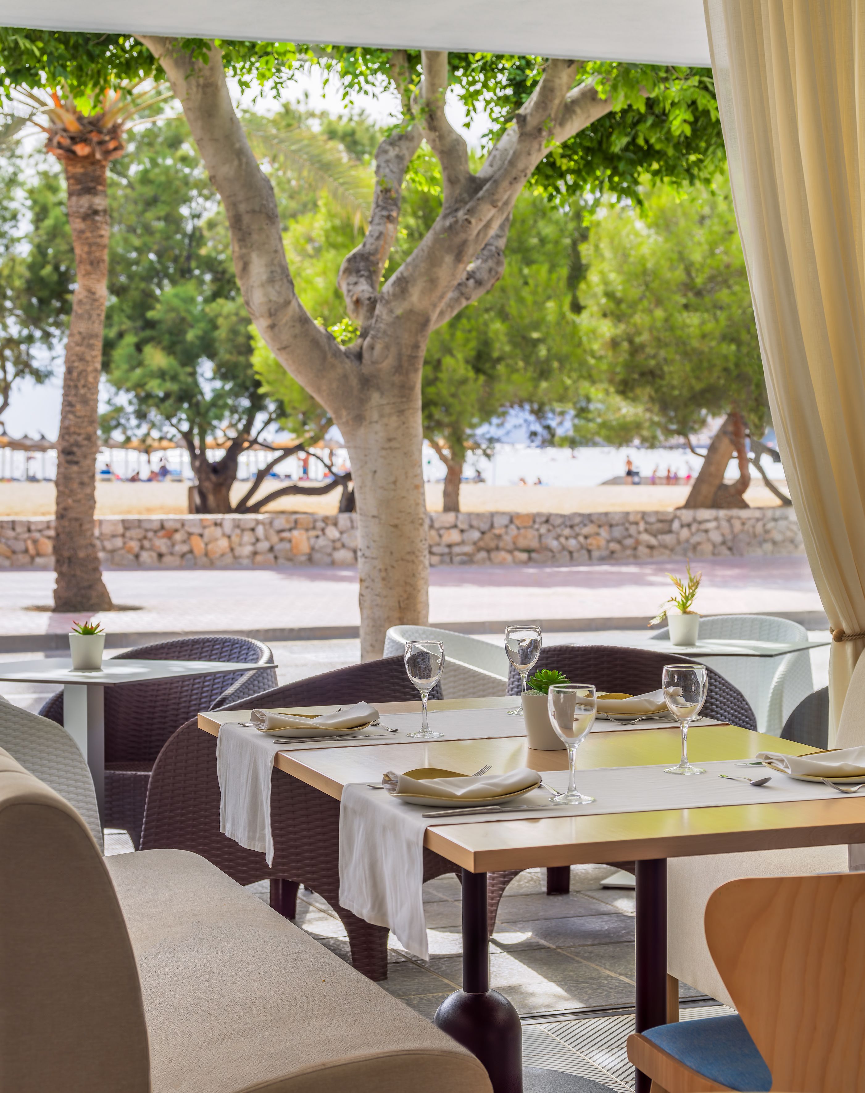 An outdoor dining area with tables set for two, surrounded by wicker chairs and a stone wall.