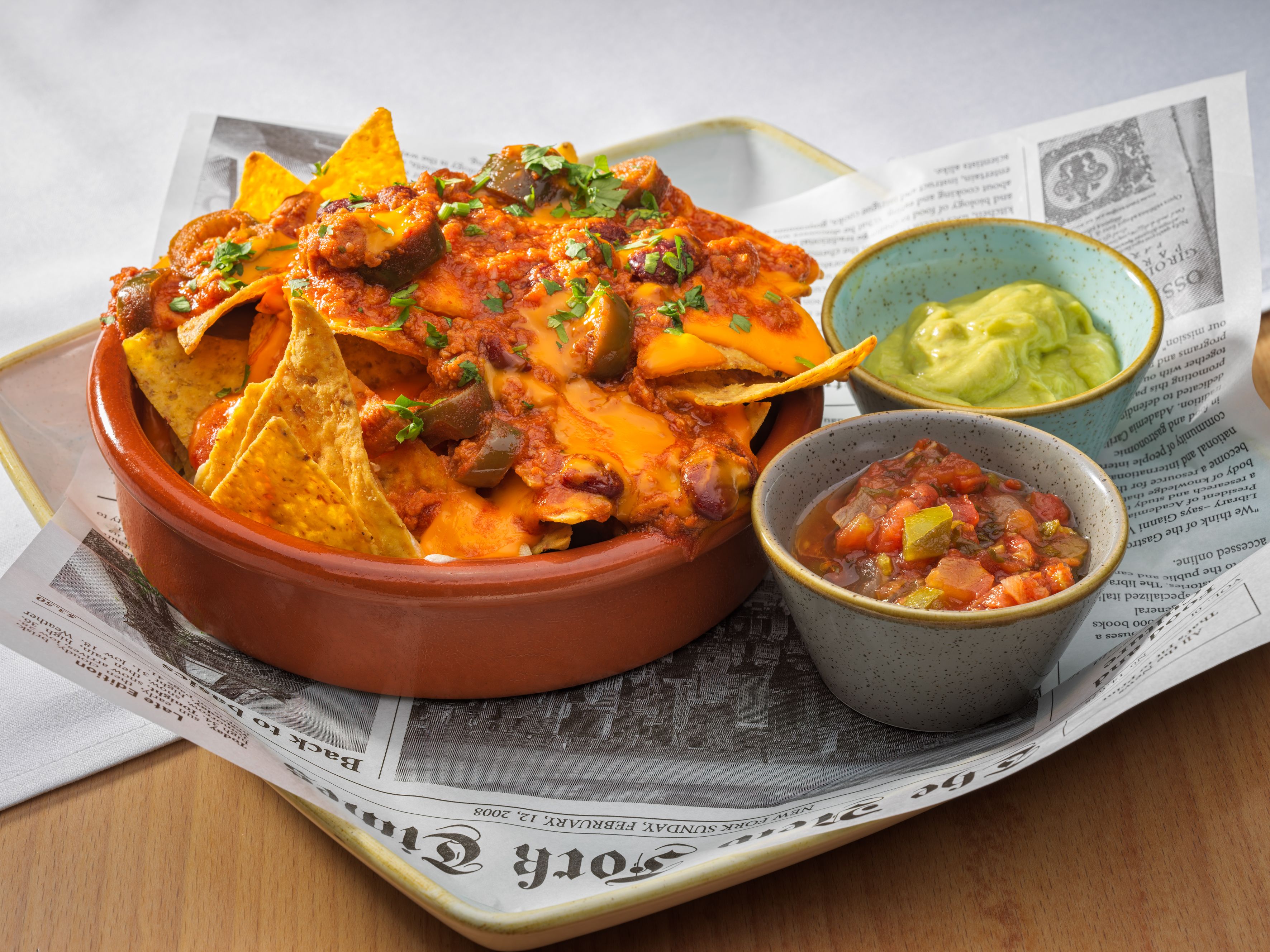 Un plato de nachos cubiertos de queso derretido y jalapeños, un tazón pequeño de guacamole y un tazón de salsa sobre un periódico.