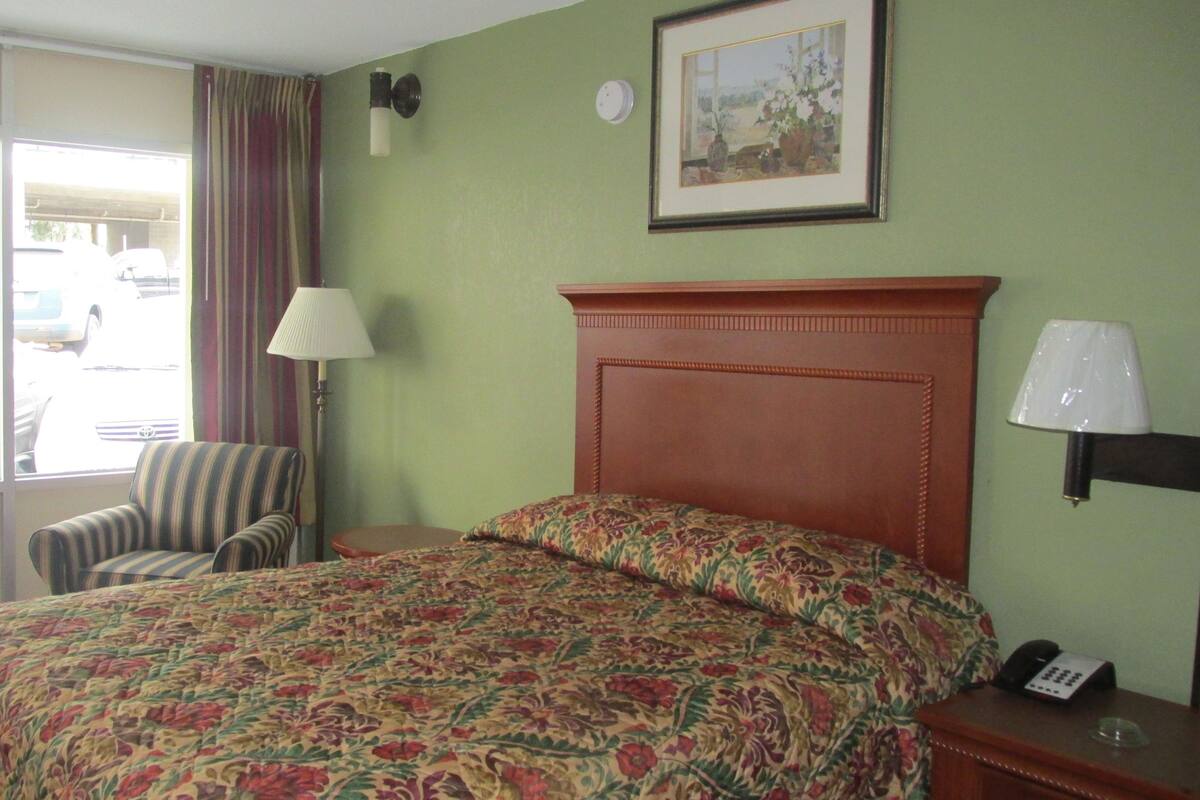 Hotelkamer met een bed, een stoel, een lamp, een schilderij en uitzicht op geparkeerde auto's.