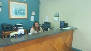 Un mostrador de recepción con computadora, impresora y varios documentos. Hay un cuadro enmarcado en la pared y un tablón de anuncios con avisos.
