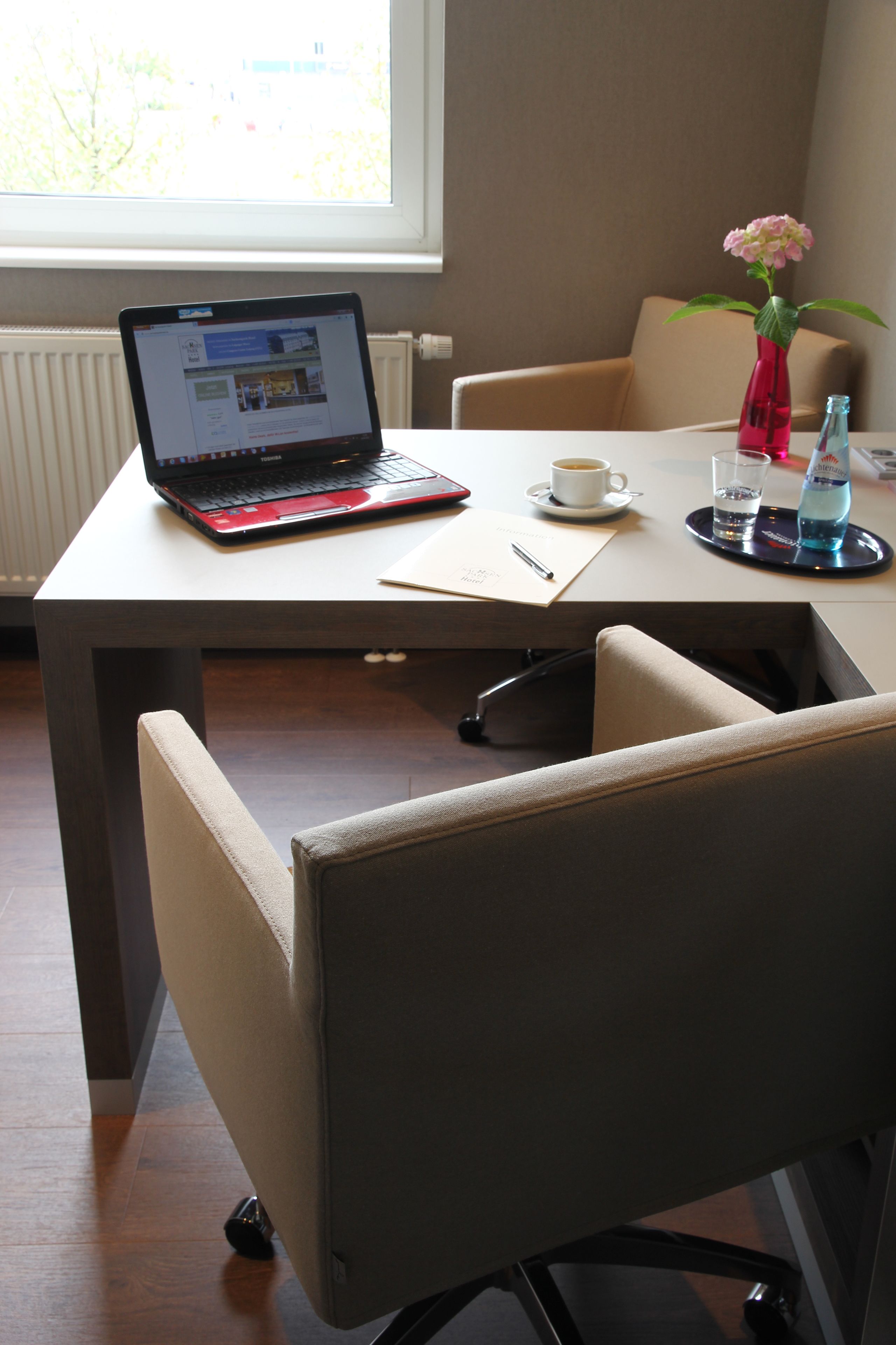 Ein Laptop auf einem Tisch, dazu eine Kaffeetasse, eine Vase mit Blumen und eine Wasserflasche.