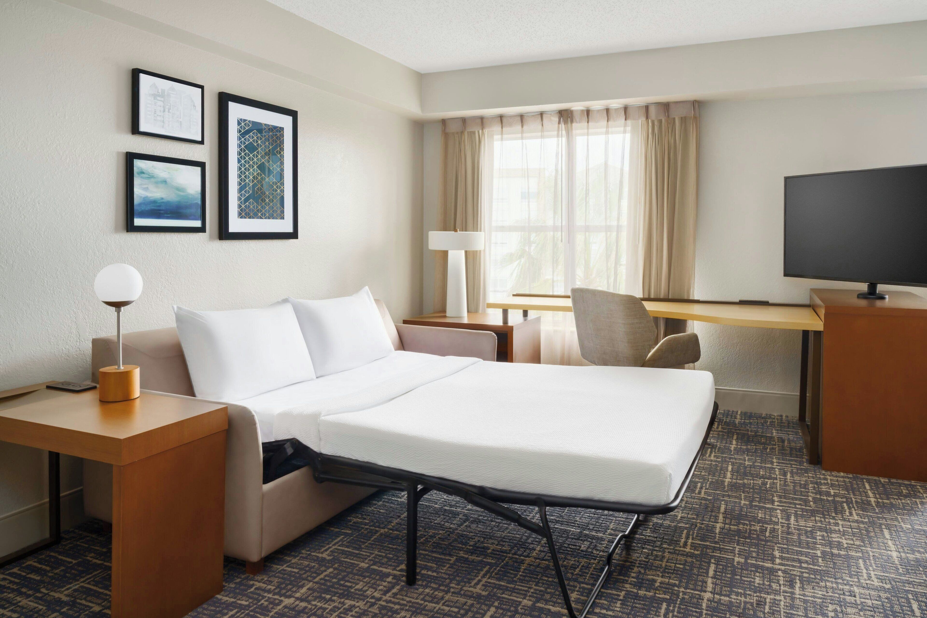 A hotel room with a bed, a desk, a chair, a TV, and framed pictures on the wall.