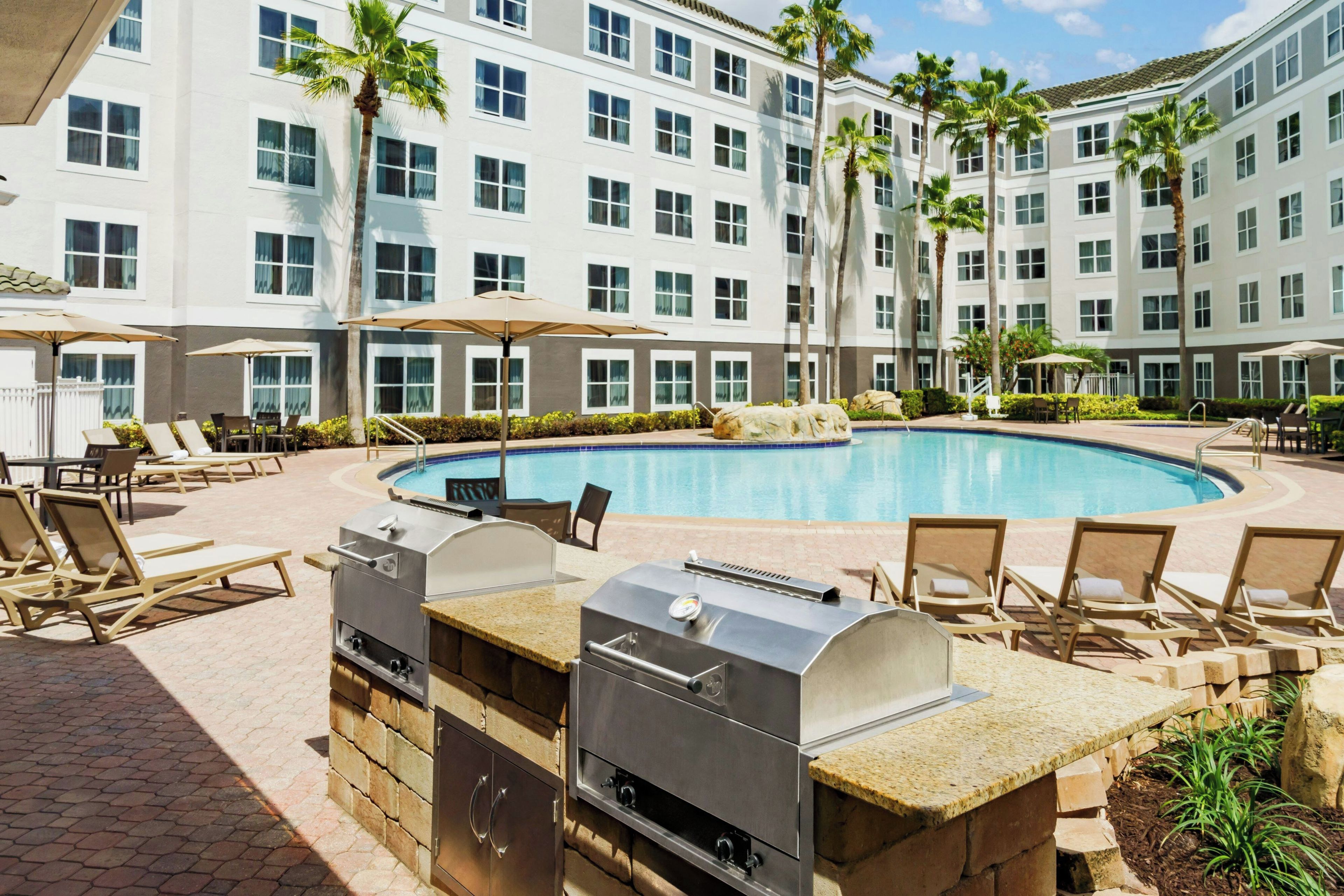 A swimming pool area with lounge chairs, a barbecue grill, and a building with multiple windows.