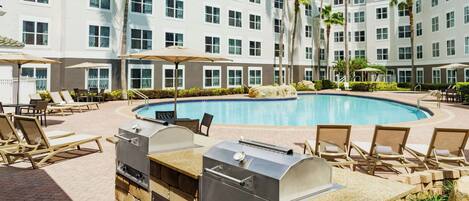 Un espace piscine avec des chaises longues, un barbecue et un bâtiment doté de nombreuses fenêtres.