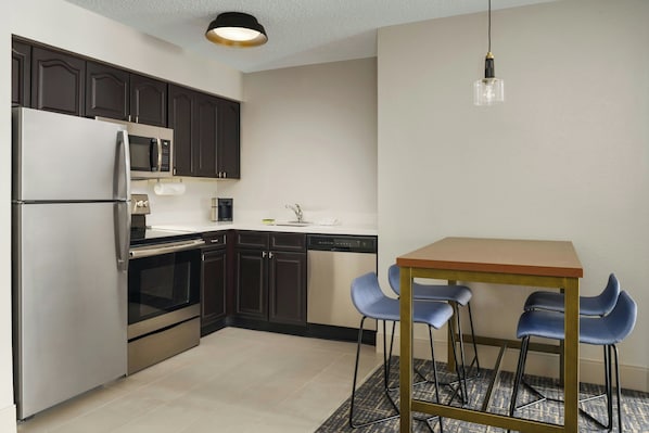 A compact kitchen with dark cabinets, stainless appliances, and a small dining area with blue chairs. - Residence Inn by Marriott Orlando Lake Buena Vista (Orlando)