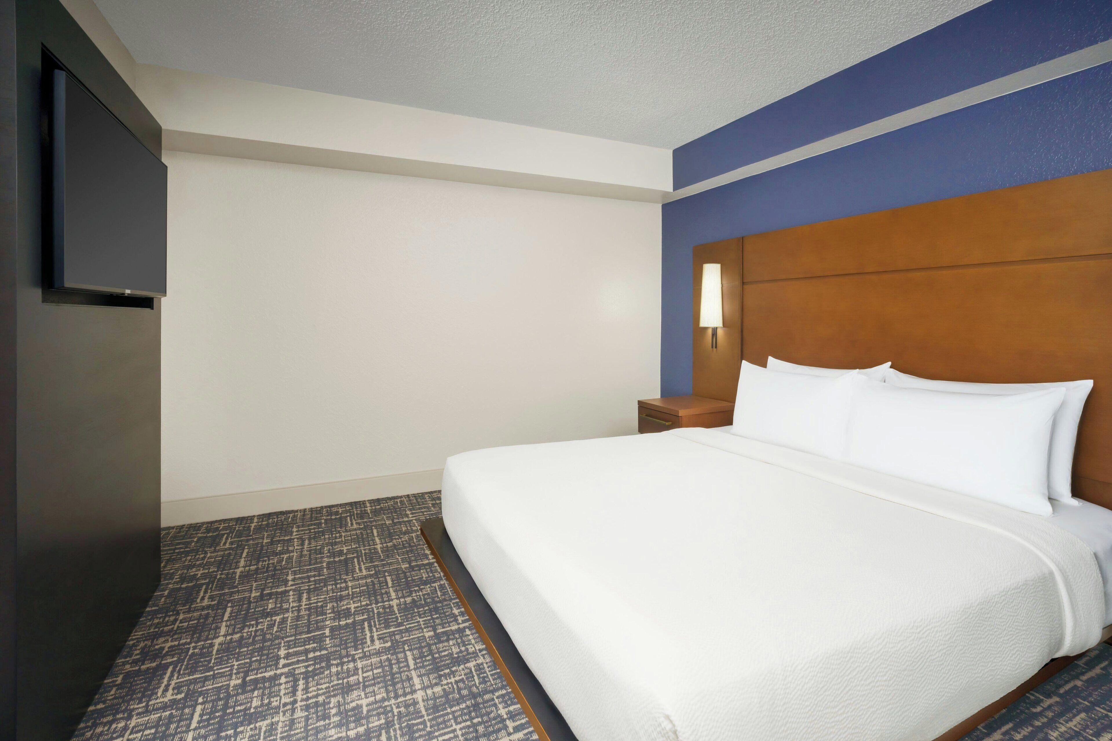 A hotel room with a large bed, a flat-screen TV mounted on the wall, a nightstand, and a patterned carpet.