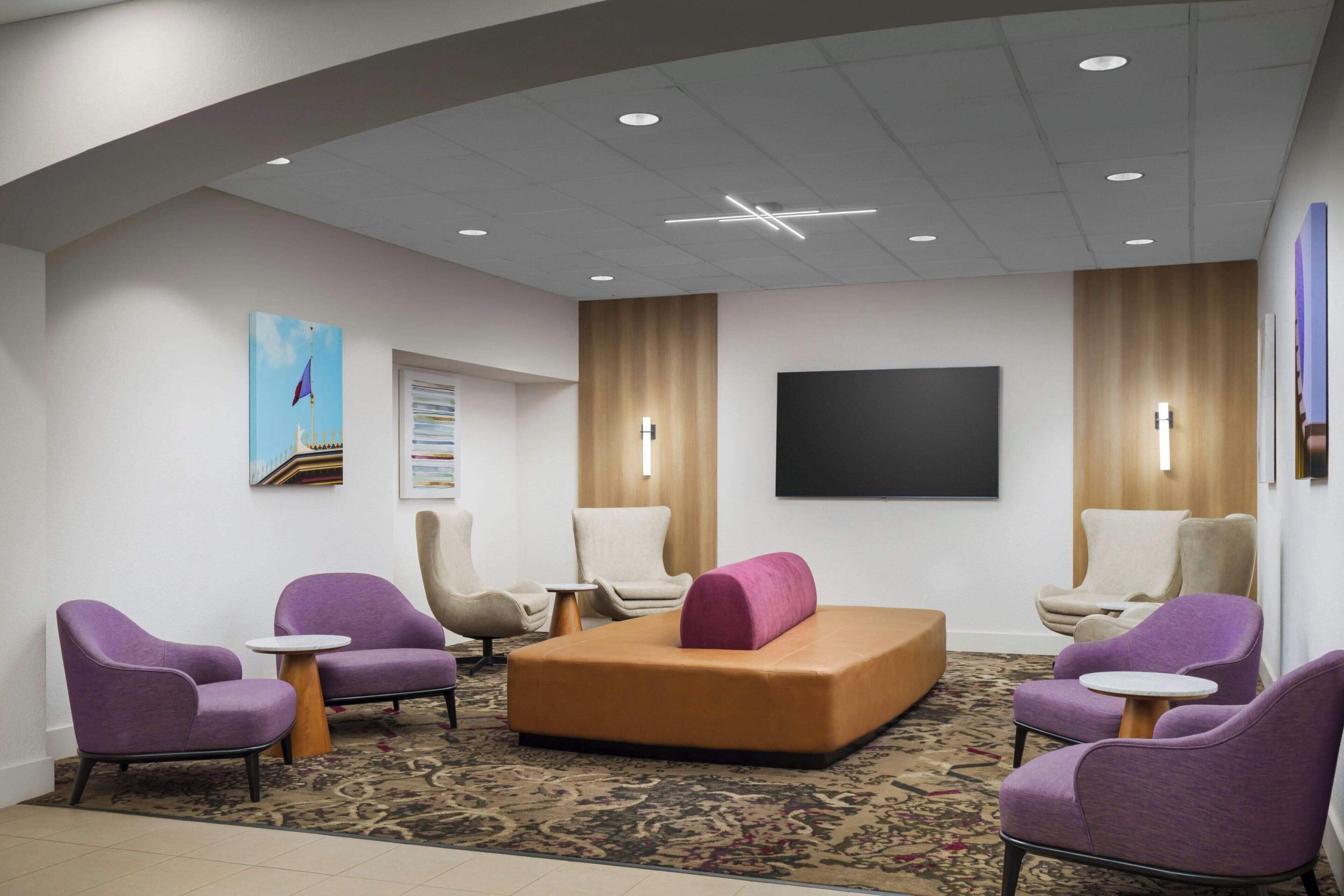 A hotel lobby with purple armchairs, a beige sofa, a flat-screen TV, and a patterned carpet.