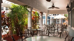 Un espace extérieur aménagé, abrité et doté de tables et de chaises rondes, entouré d’une végétation tropicale luxuriante et de colonnes.