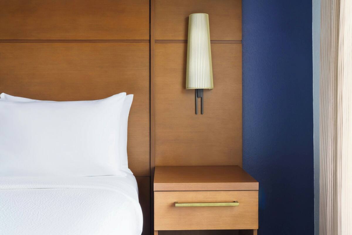 Ein ordentlich gemachtes Bett mit weißer Bettwäsche, ein Nachttisch aus Holz mit einer Schublade und eine an der Wand befestigte Lampe.