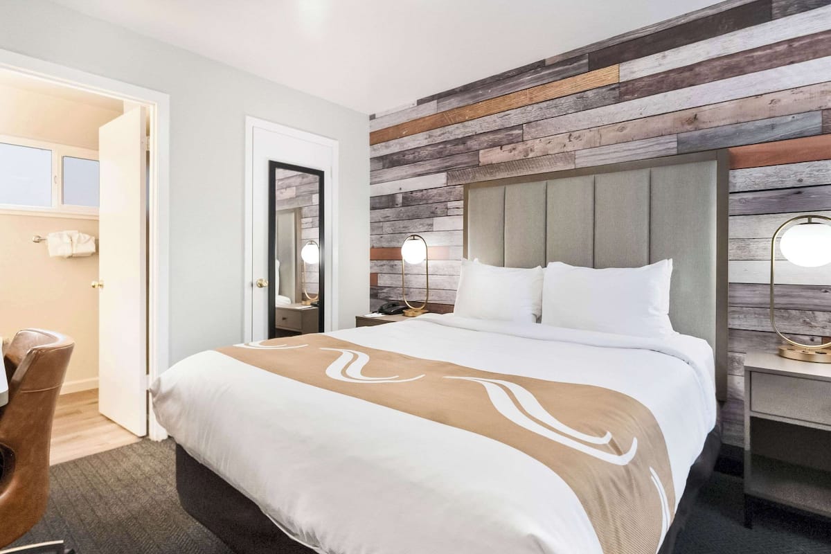 A hotel room with a large bed, a wooden accent wall, a desk with a lamp, and a chair.