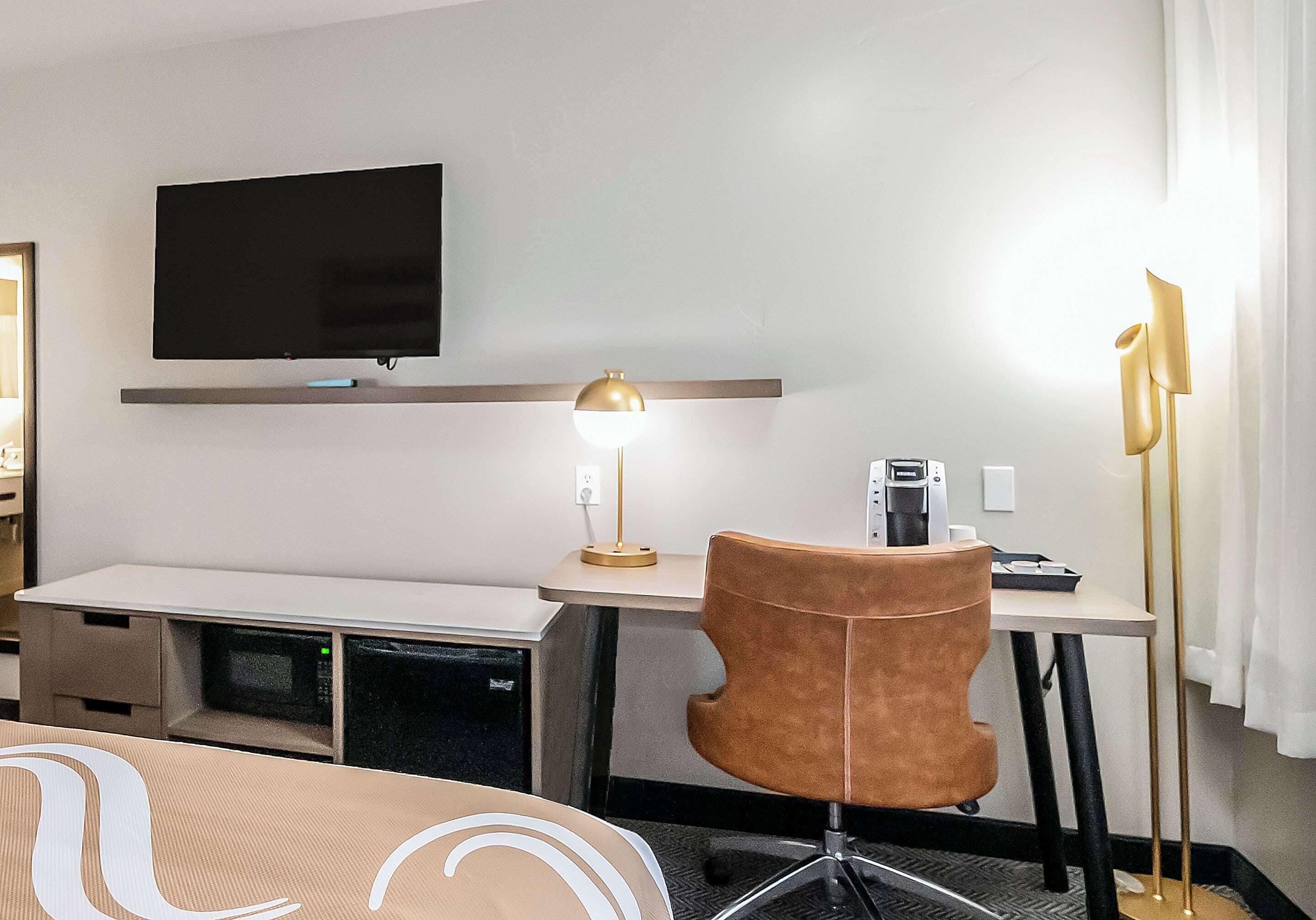 A hotel room with a flat-screen TV mounted on the wall, a desk with a lamp, a chair, and a coffee machine.