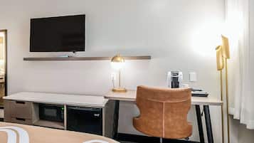 A hotel room with a flat-screen TV mounted on the wall, a desk with a lamp, a chair, and a coffee machine.