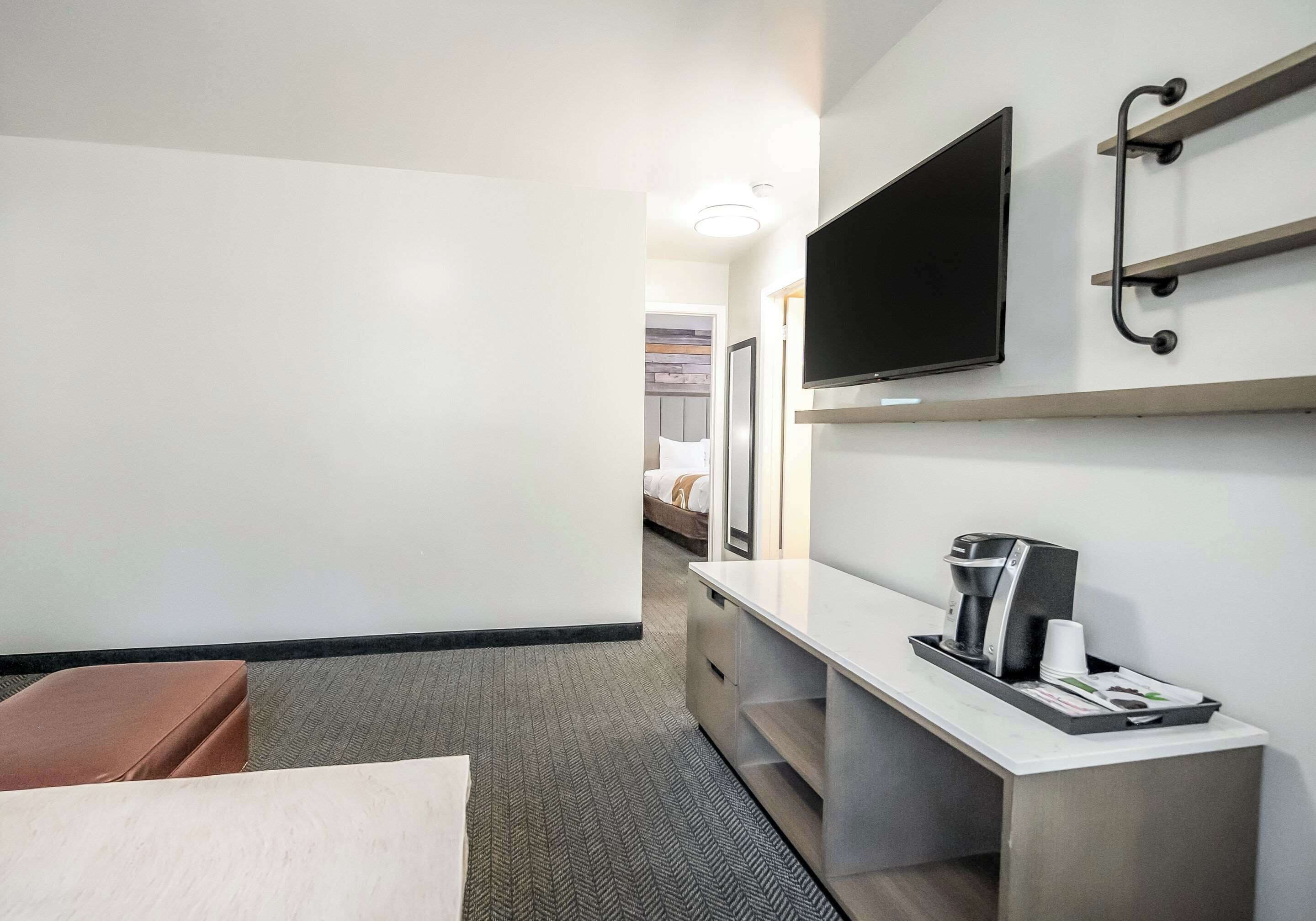 A hotel room with a bed, a desk with a coffee maker, a TV mounted on the wall, and a small seating area.