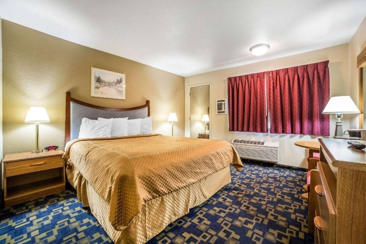 A hotel room with a bed, bedside lamps, a desk, a chair, a window with curtains, and a painting on the wall.