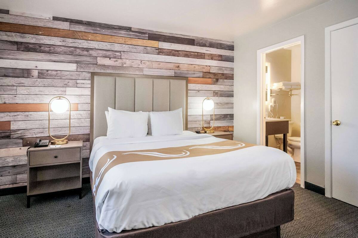 A hotel room with a wooden headboard, a large bed with white bedding, two bedside lamps, a nightstand, and a bathroom visible through an open door.