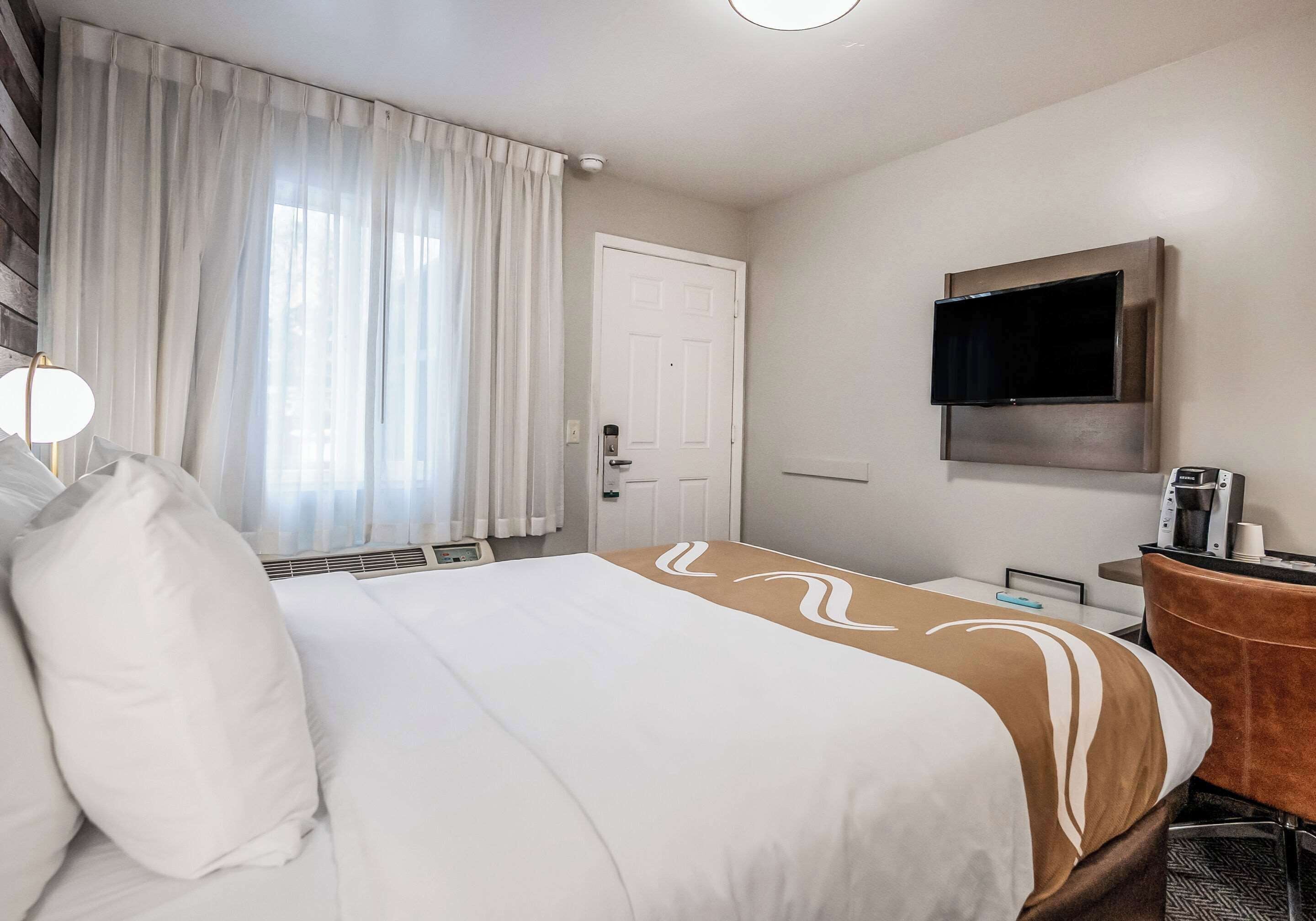 A hotel room with a large bed, a flat-screen TV mounted on the wall, a desk with a coffee machine, and a door with a handle.