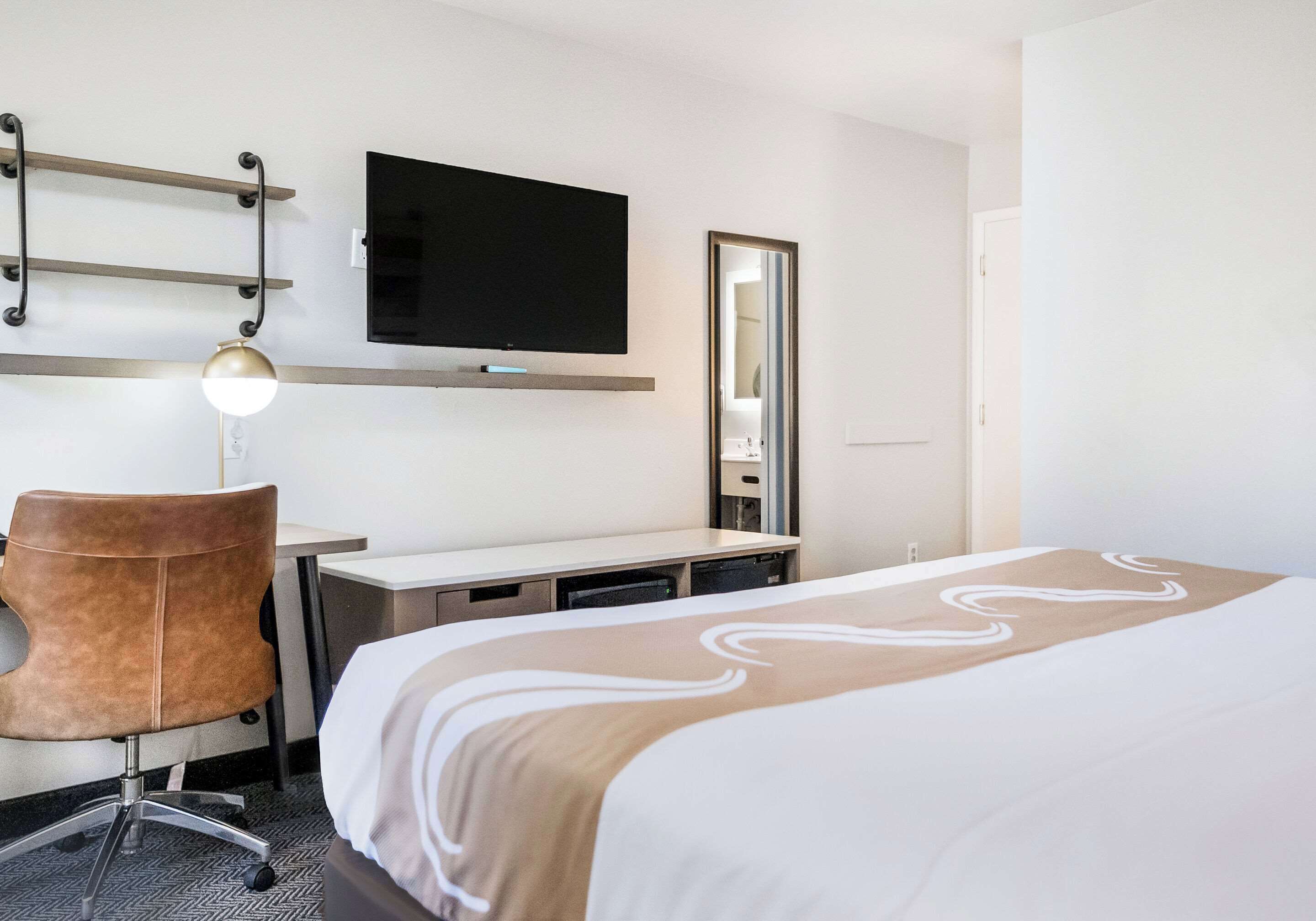 A hotel room with a bed, a desk, a chair, a TV, and a mirror.