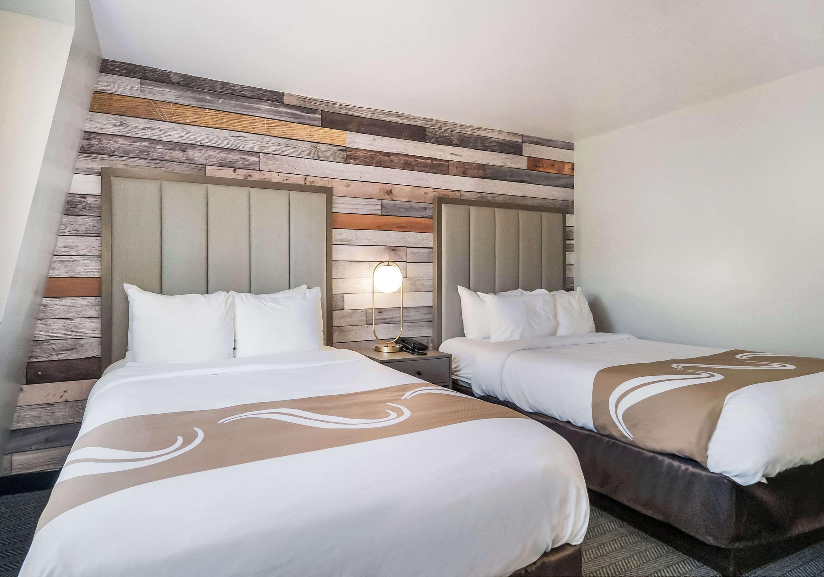 A hotel room with two beds, a wooden wall panel, a nightstand with a lamp, and a phone.