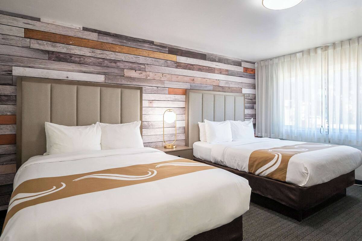A hotel room with two beds, a wooden wall panel design, a large window with sheer curtains, and a carpeted floor.