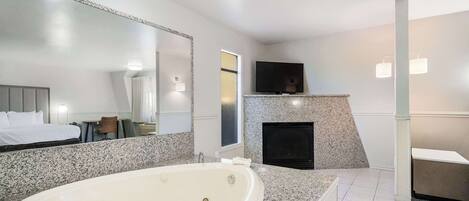 A modern hotel bathroom with a large freestanding bathtub, a mirror, a fireplace, and a television mounted on the wall.
