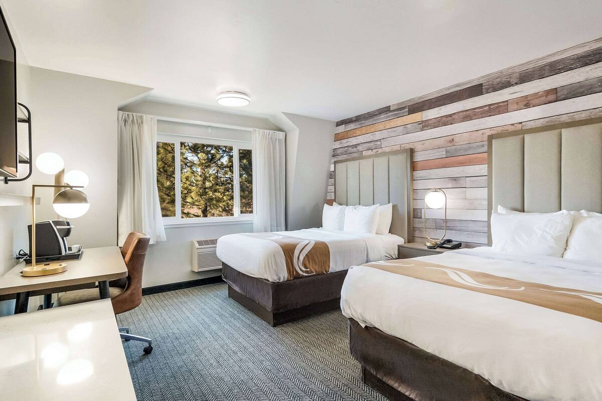 A hotel room with two beds, a desk with a lamp, a chair, and a window with curtains.