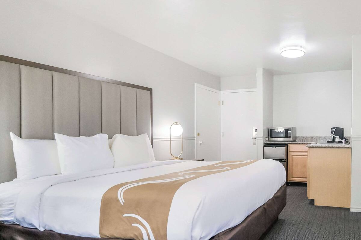 A hotel room with a large bed, a bedside table with a lamp, a microwave, and a small kitchenette.