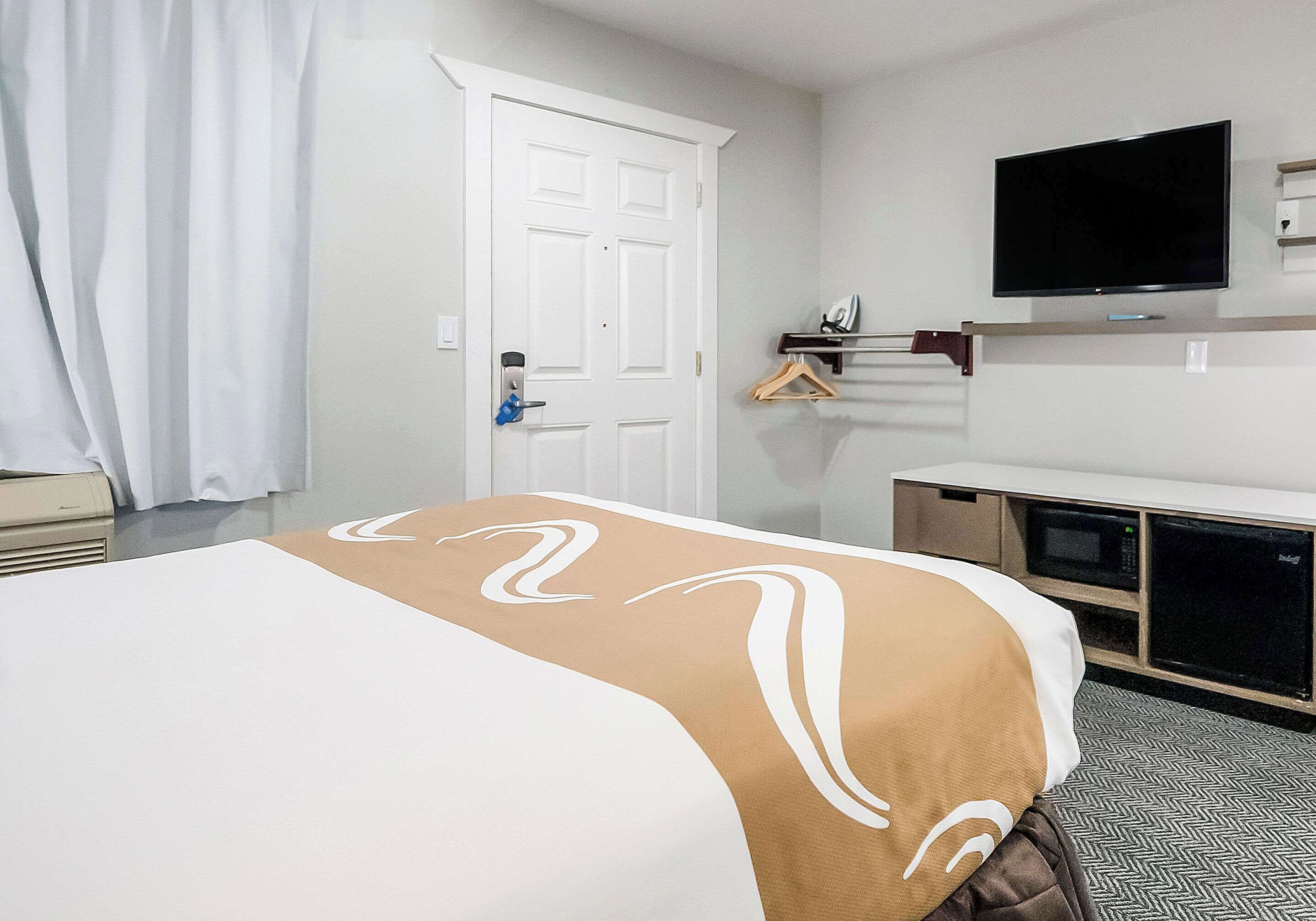 A hotel room with a bed, a television mounted on the wall, a microwave, and a small shelf with a hanger.