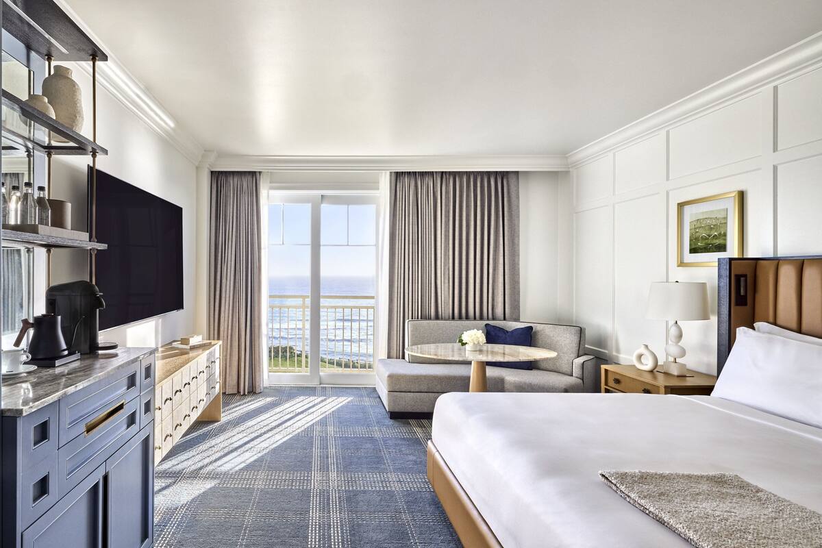 A hotel room with a bed, a sofa, a TV, a coffee machine, a desk, and a view of the outdoors.