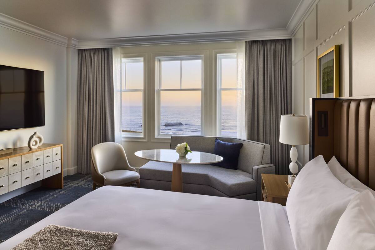 A hotel room with a large bed, a sofa, a round table with a vase, a TV, and a view of the ocean.