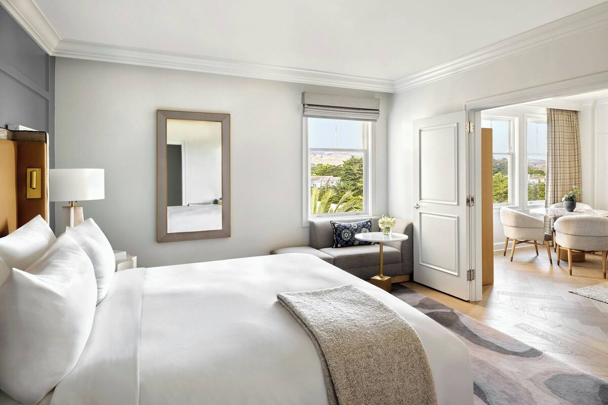 A hotel room with a large bed, a seating area with a table, a window with a view, and a door leading to another room.