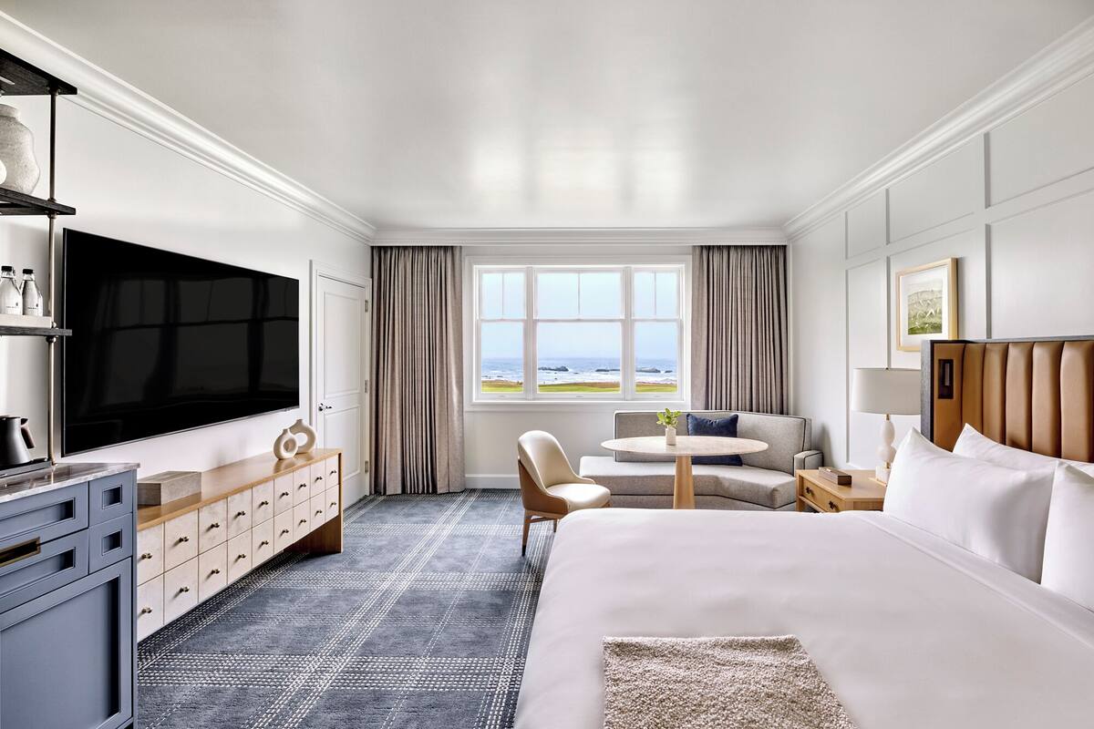 A hotel room with a large bed, a TV, a desk with a chair, a window with a view of the ocean, and a minibar.
