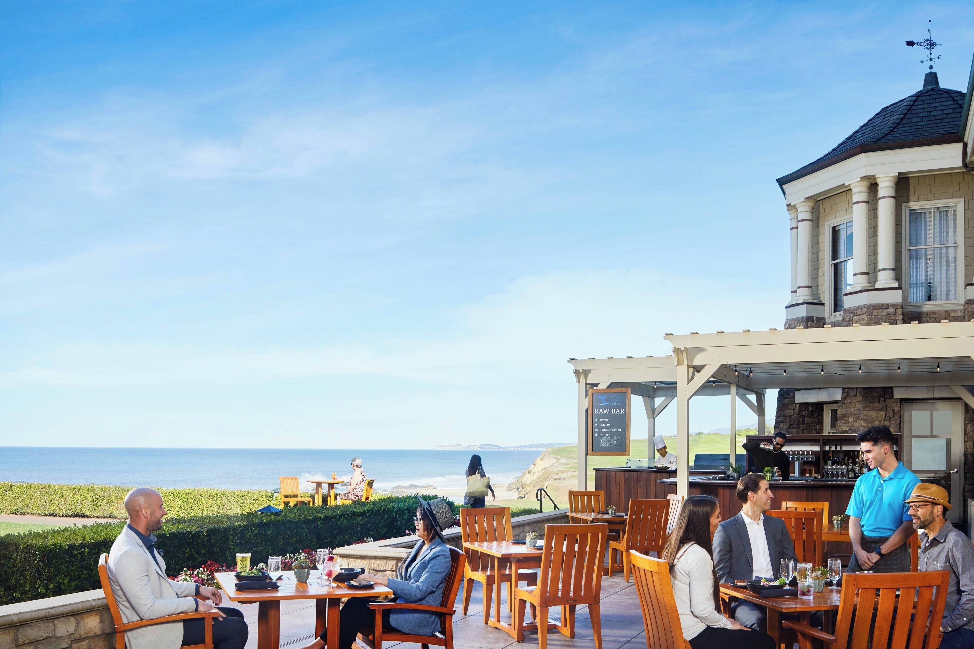 A coastal restaurant with outdoor seating overlooking the ocean, featuring a bar and a view of the beach.