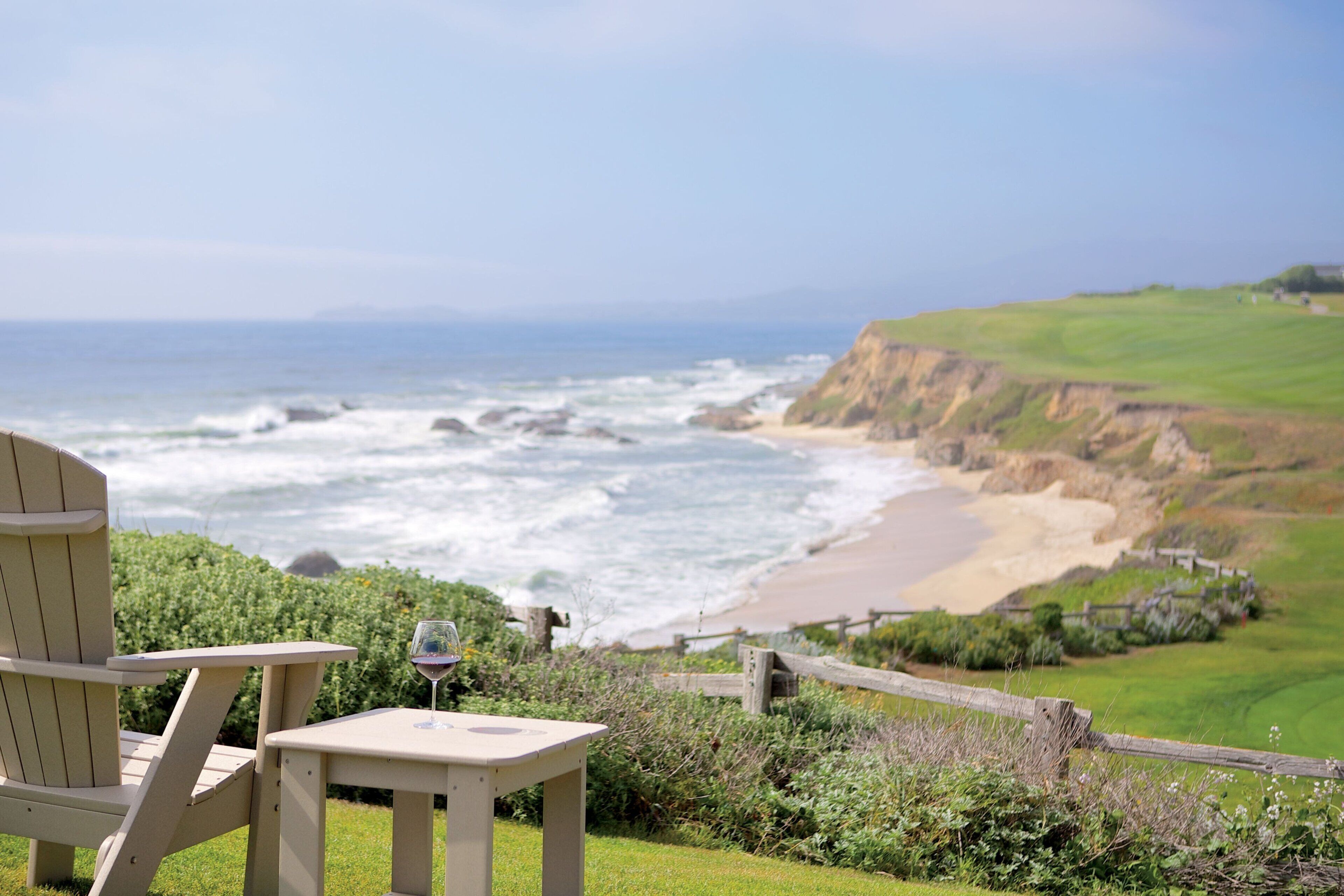 A beach view with a wooden chair and table, a glass of wine, and a grassy area with a wooden fence.