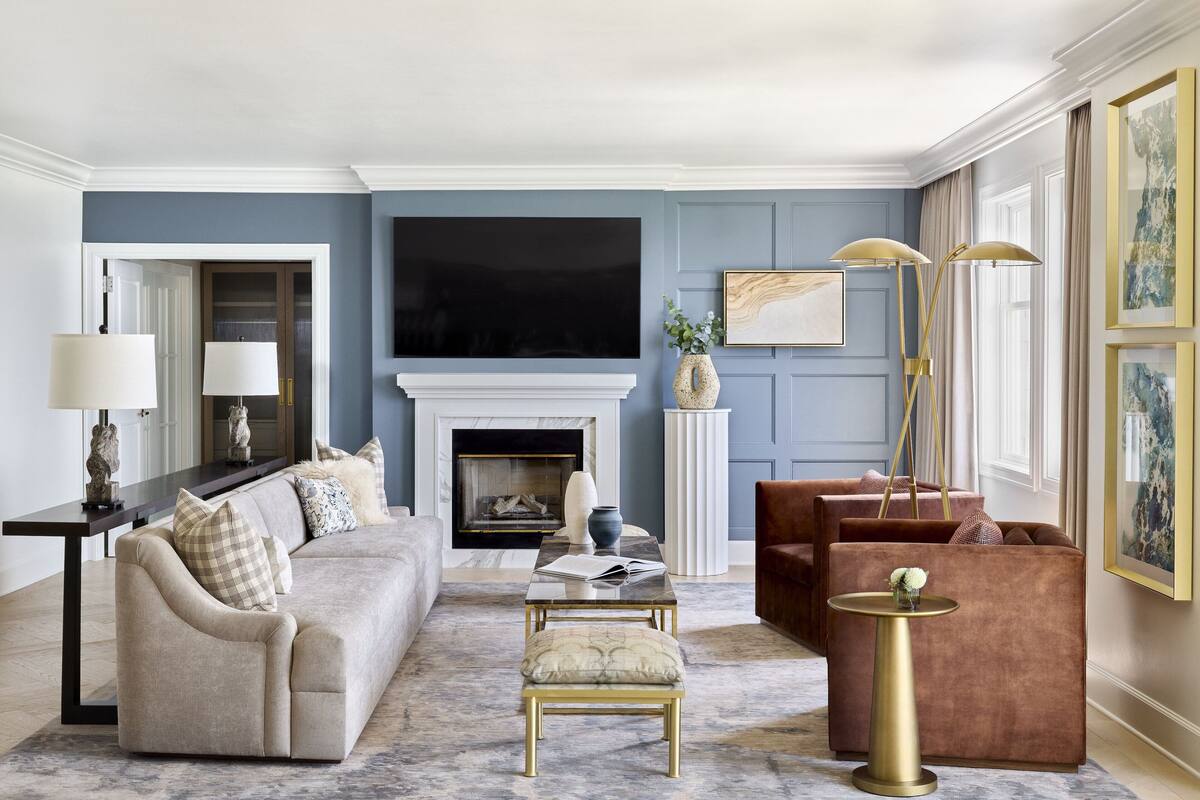 A living room with a sofa, armchair, and ottoman, a fireplace, a TV, and a side table with a lamp.