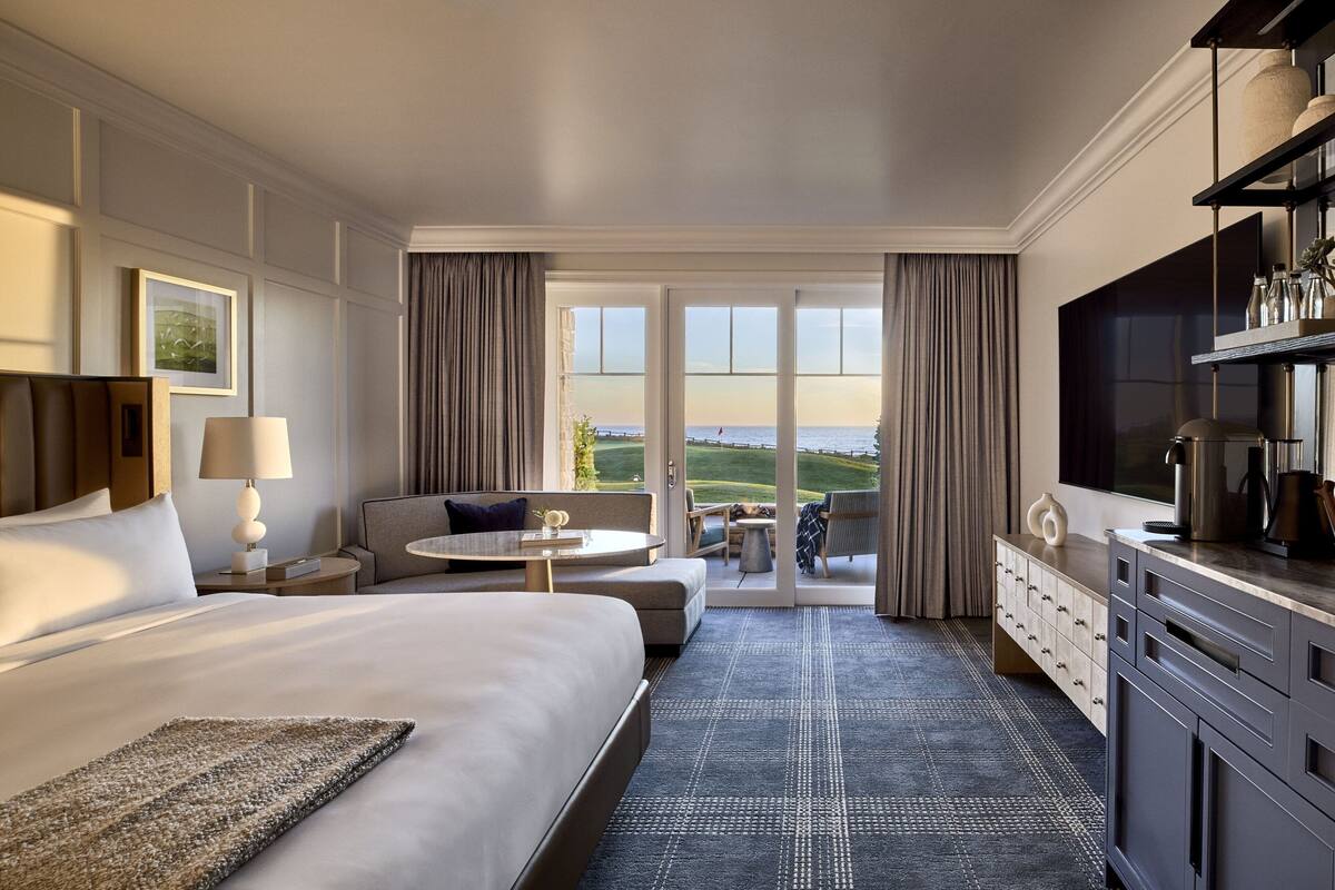 A hotel room with a large bed, a sofa, a dining table, a TV, and a view of the ocean.