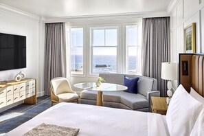 A hotel room with a bed, a sofa, a round table, a TV, and a view of the ocean.