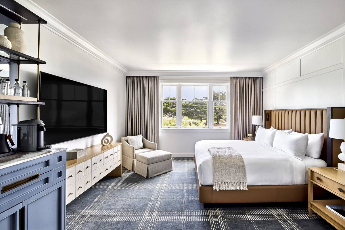 A hotel room with a large bed, a TV, a desk with a lamp, a chair, and a view of the outdoors.