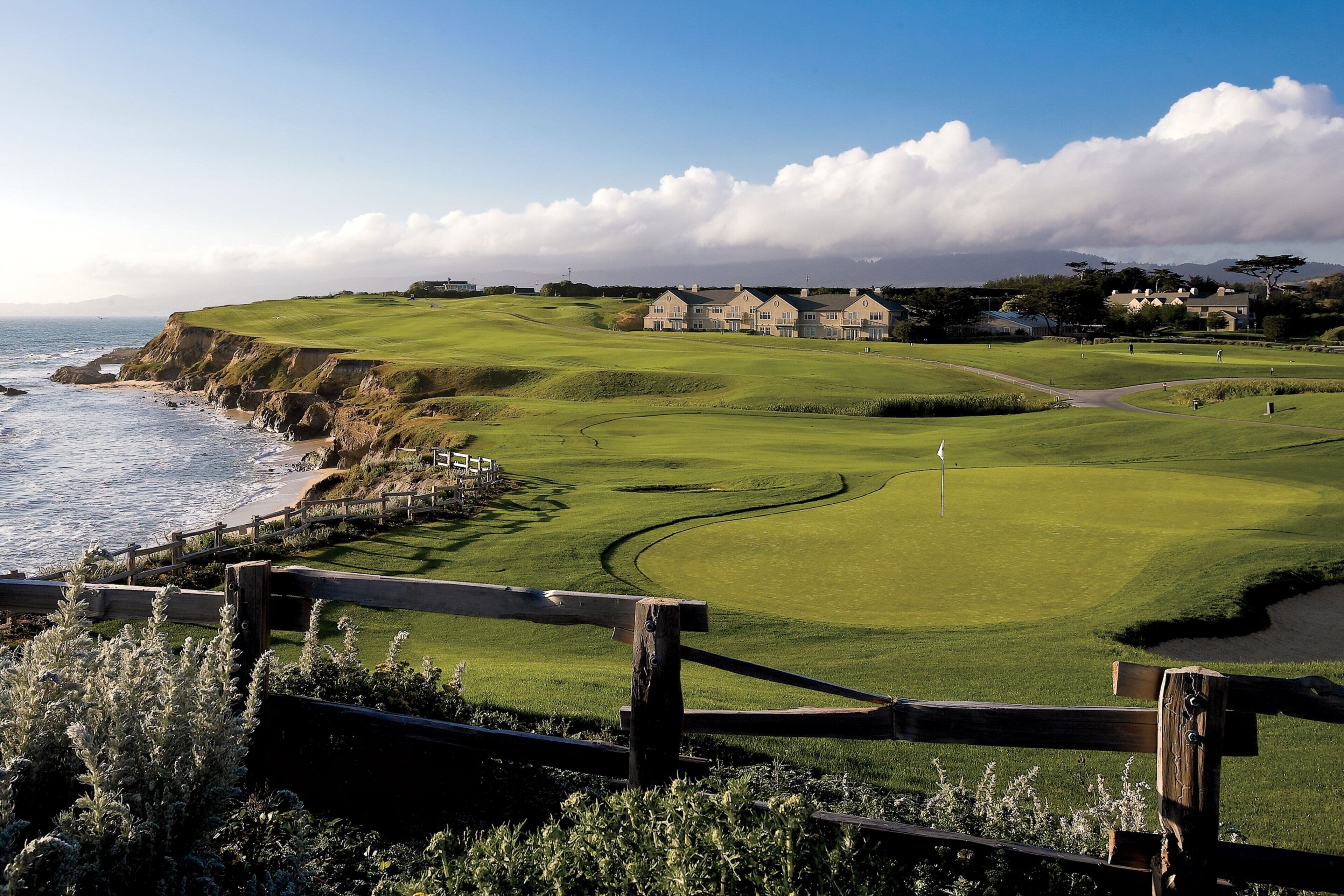 A coastal golf course with a wooden fence in the foreground, a clear sky, and houses in the background.