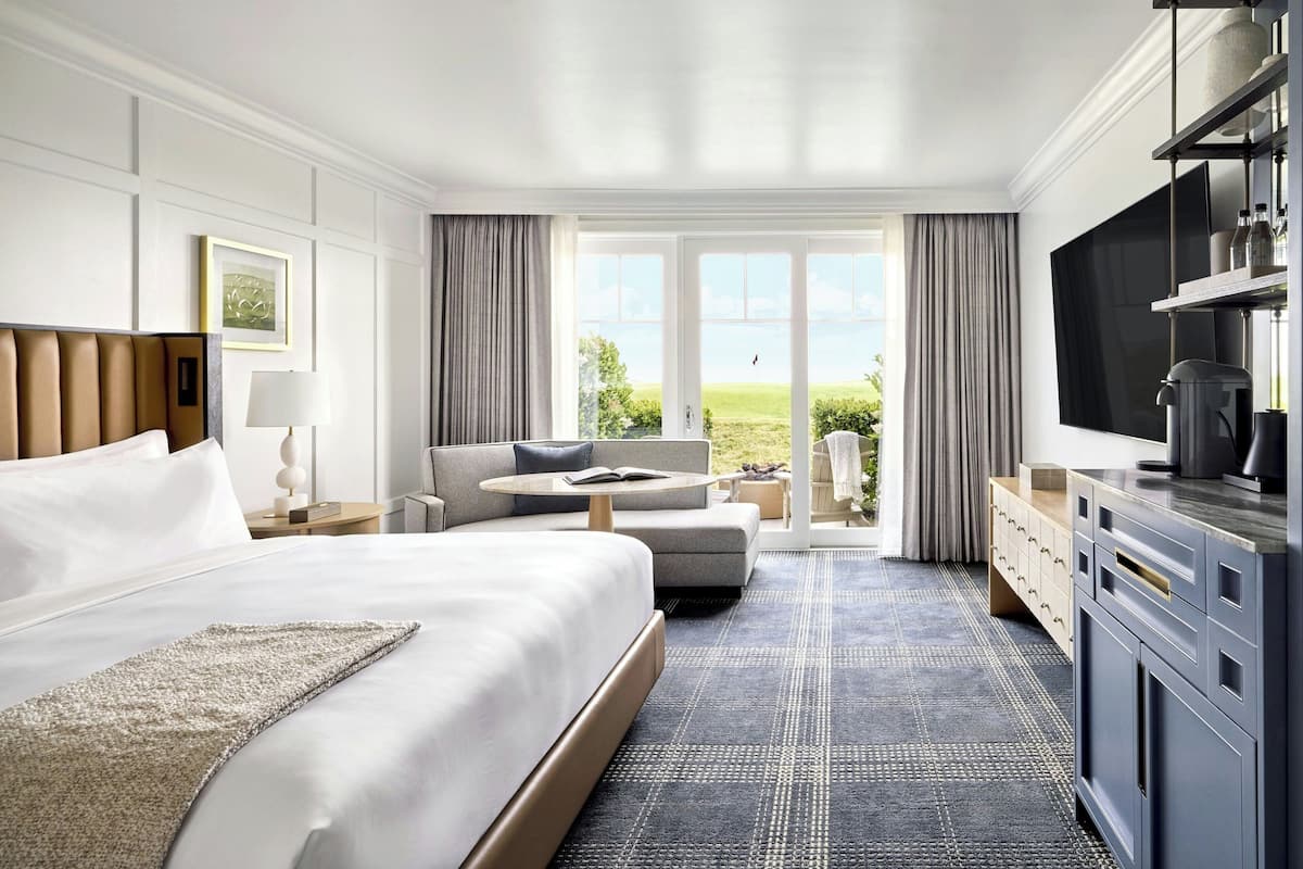 A modern hotel room with a large bed, a sofa, a dining area, and a view of the outdoors.