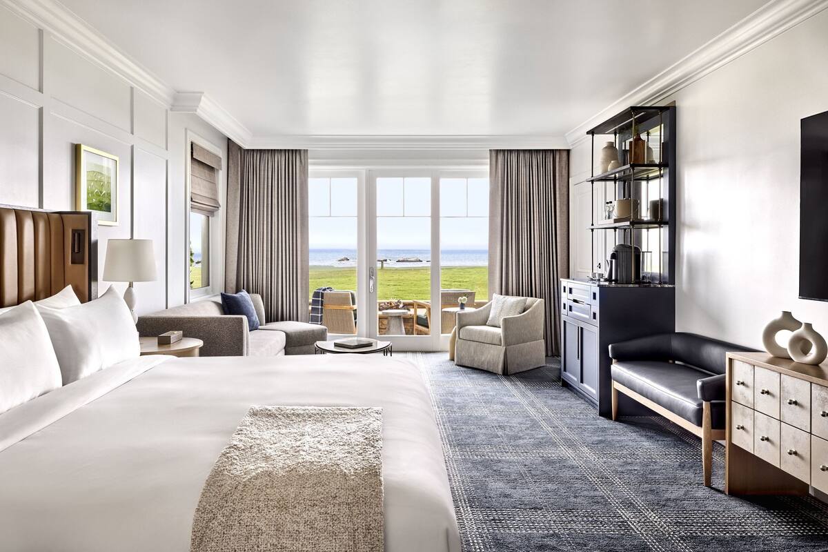 A hotel room with a large bed, a sofa, a chair, a small table, and a view of the sea.