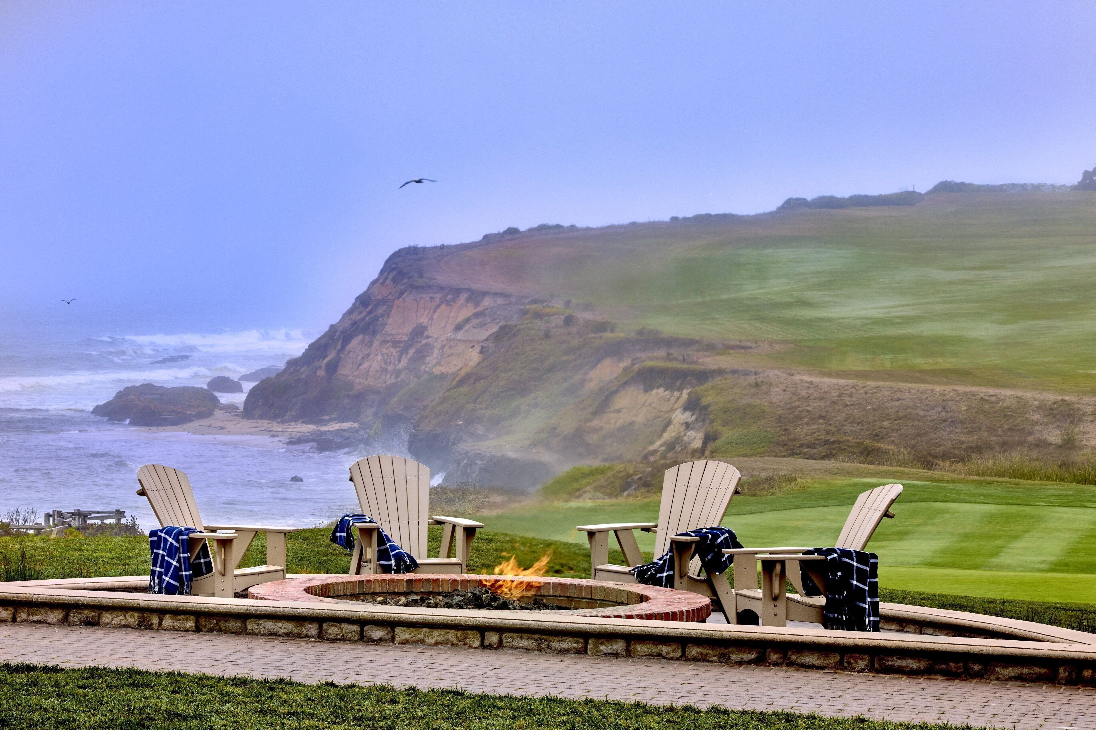 A patio with four Adirondack chairs and a fire pit overlooking a coastal landscape with cliffs and a golf course.
