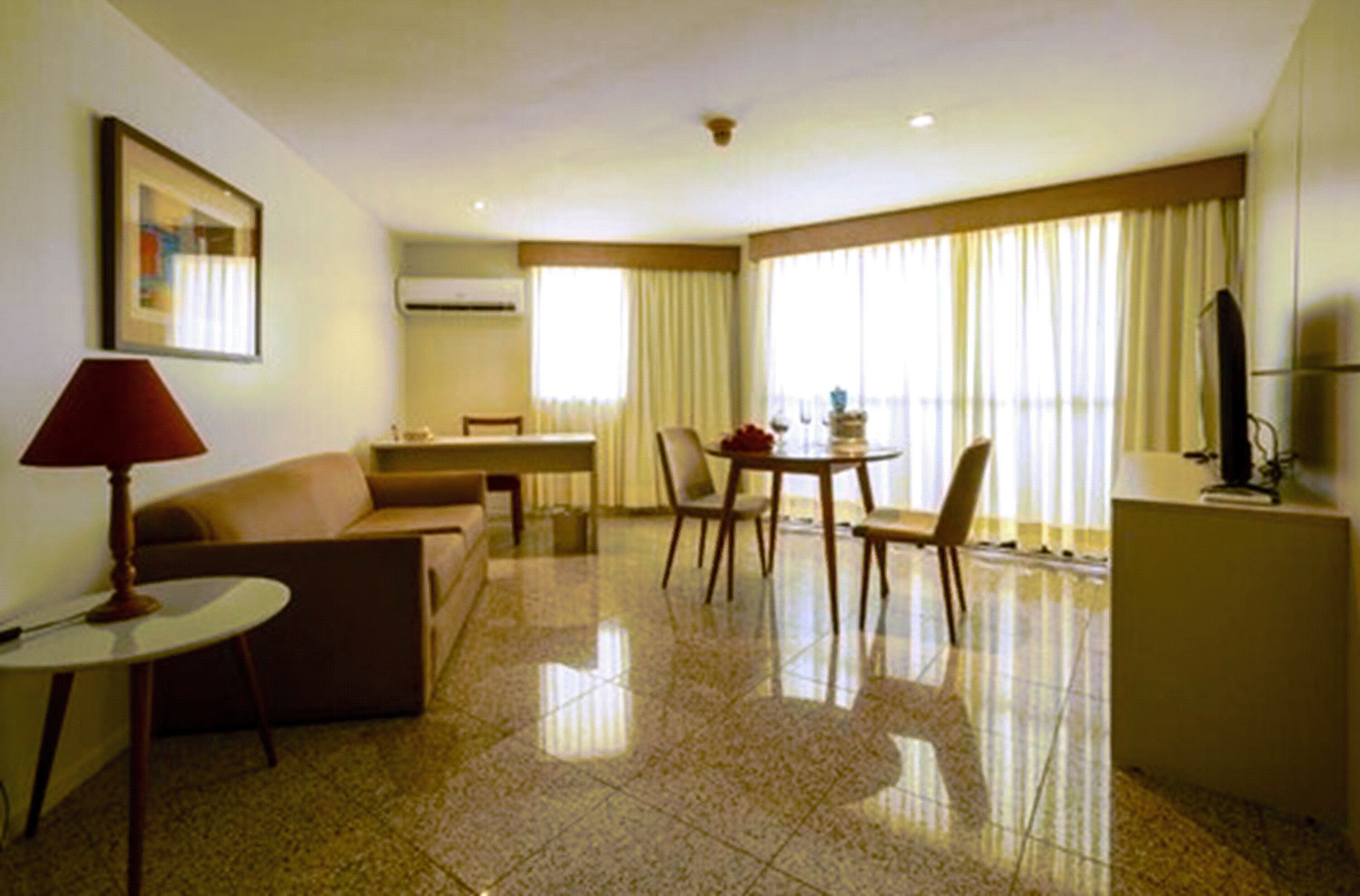 A hotel room with a sofa, a dining table with chairs, a TV, and a window with curtains.