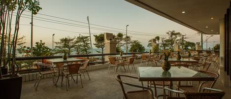 An outdoor cafe area with tables and wicker chairs, surrounded by potted plants and overlooking a cityscape.
