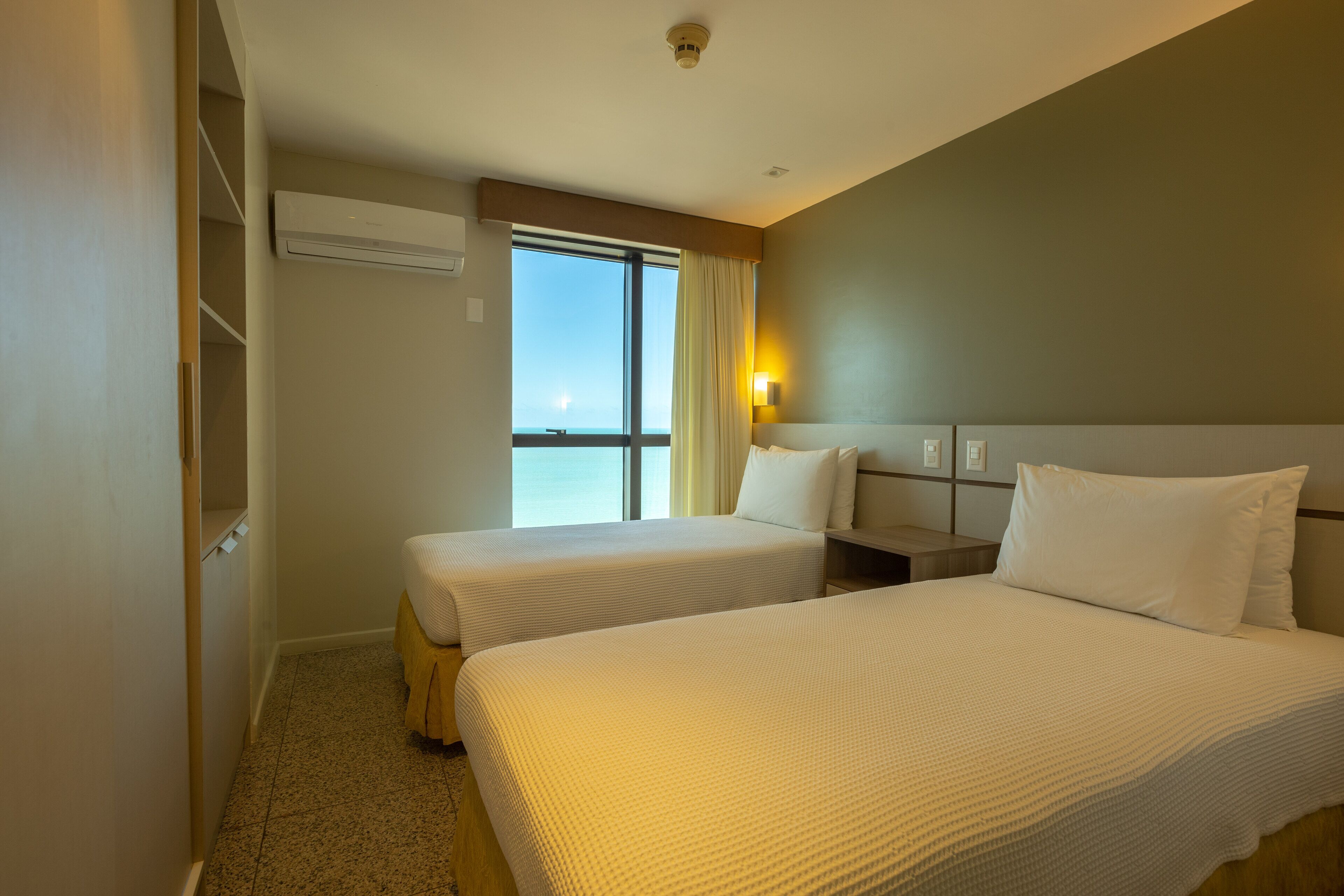 A hotel room with two beds, a large window, an air conditioning unit, and a nightstand with a lamp.