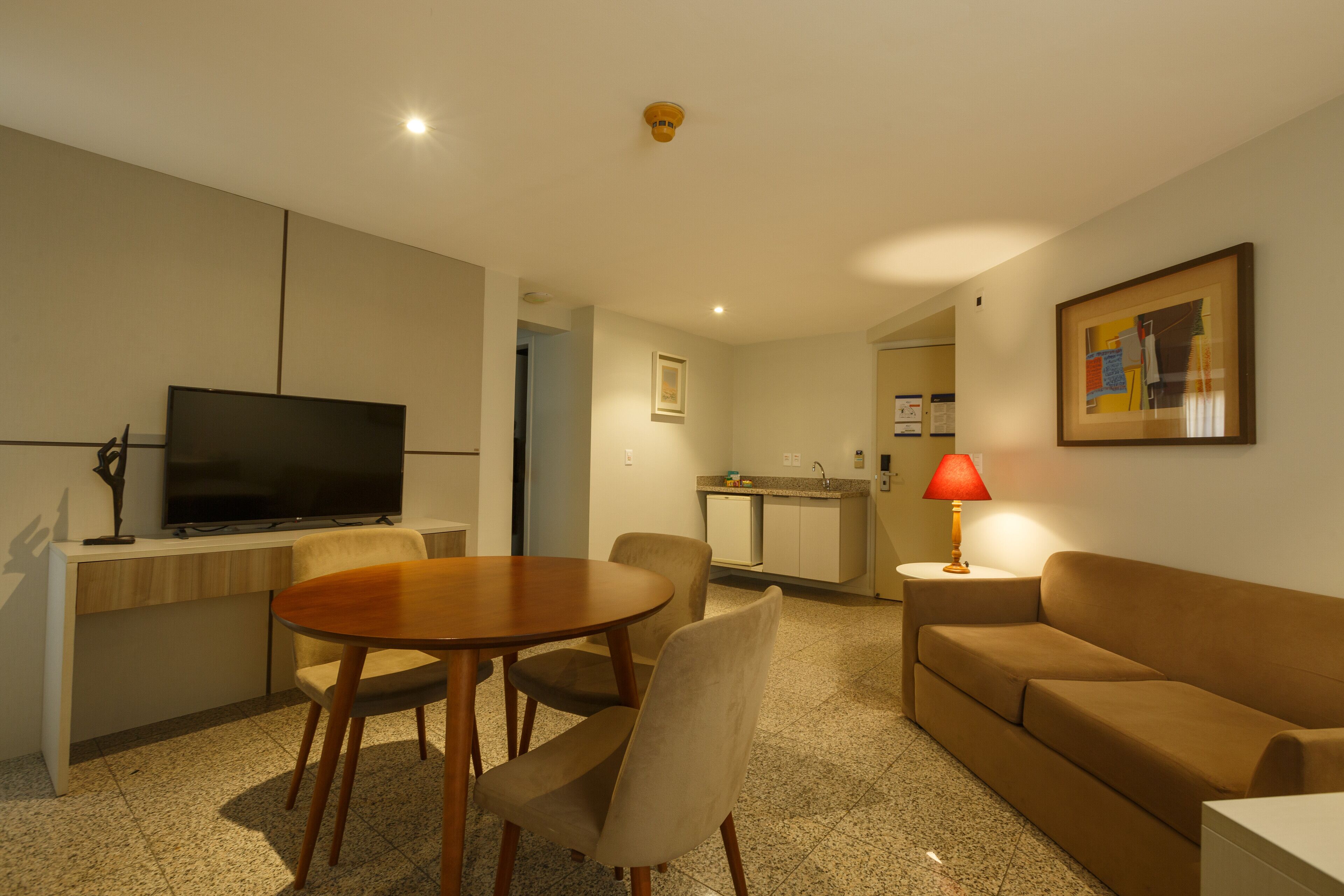 A modern hotel room with a dining area, a sofa, a TV, and a kitchen area.