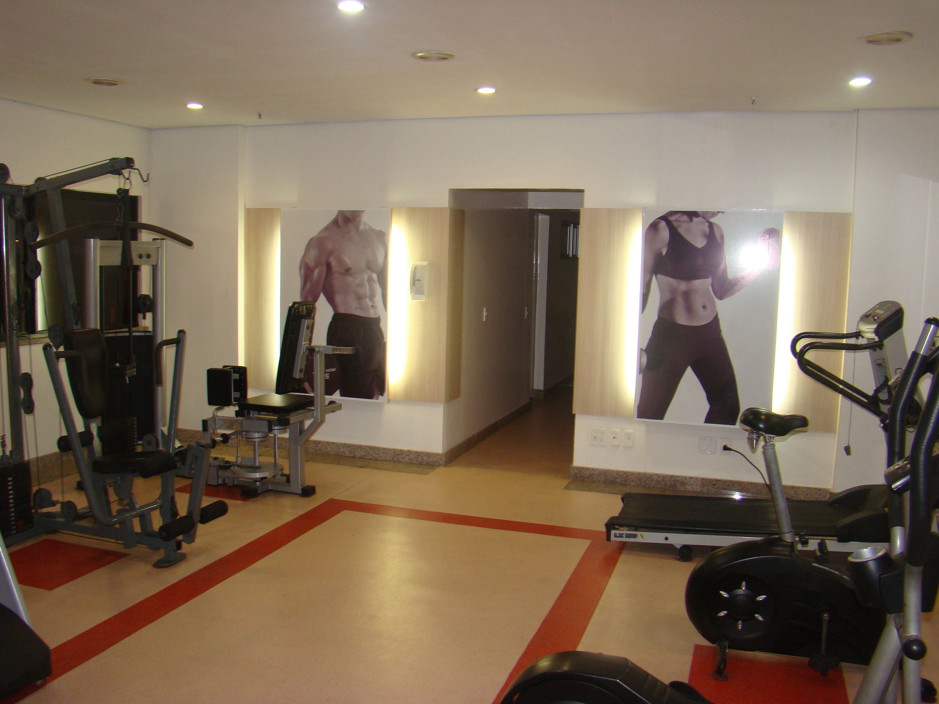 Ginásio com equipamentos de exercícios, esteira e espelhos refletindo o espaço.