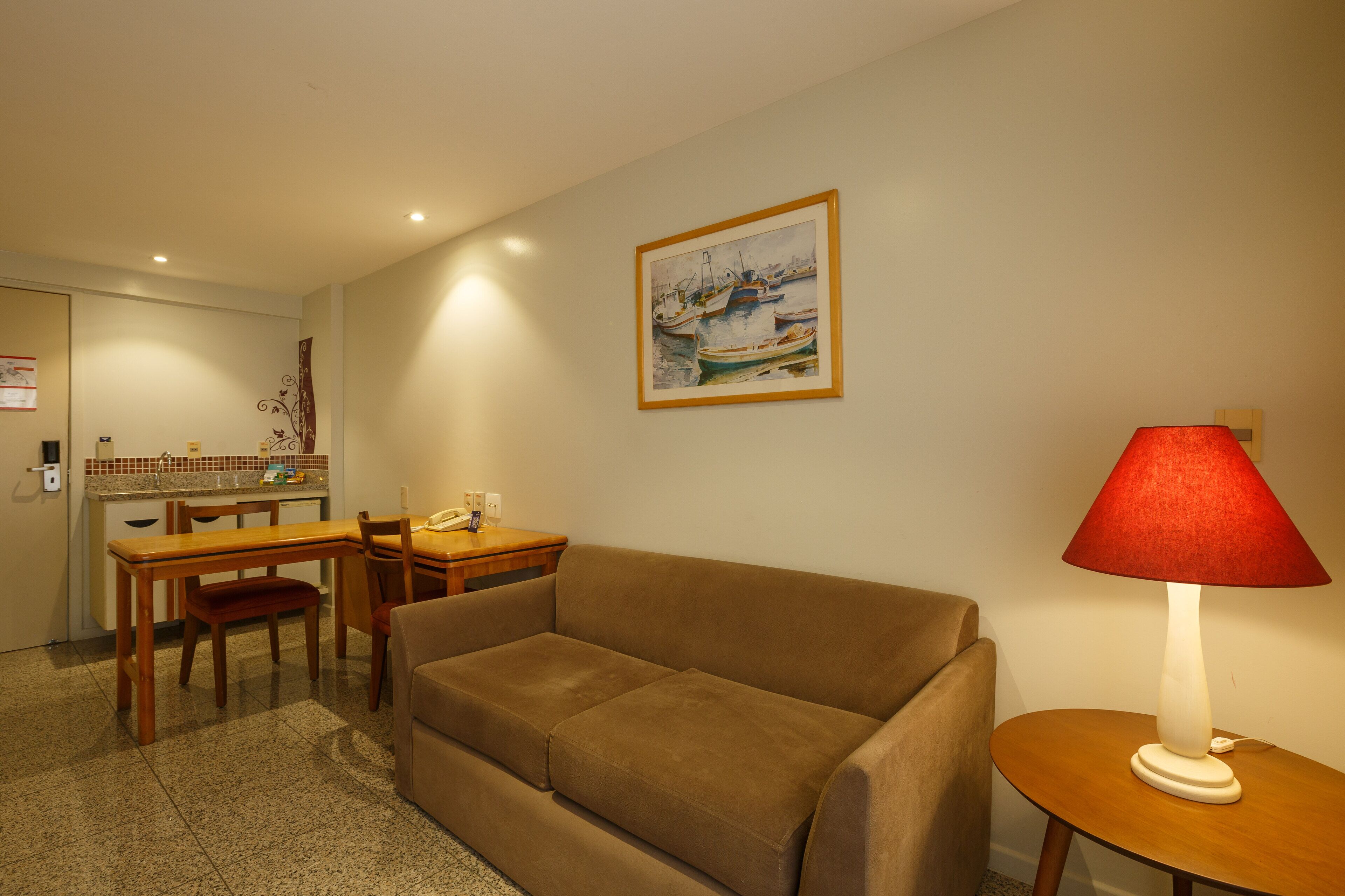 A living room with a sofa, a dining table, and chairs. There is a painting on the wall and a lamp on a side table.