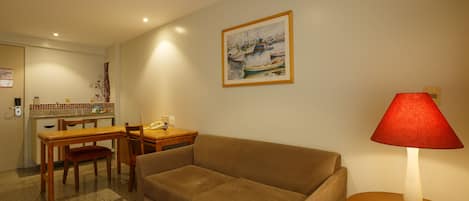 A living room with a sofa, a dining table, and chairs. There is a painting on the wall and a lamp on a side table.